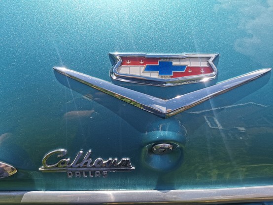 The centre of the boot / trunk lid of a Marine Blue Impala. In the centre of the image is a Chevrolet badge above a chrome V and key lock.
Just below and to the left of the lock is a Calhoun Dallas badge.