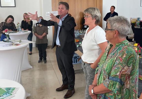 Im Vordergrund sind drei Personen zu sehen, im Hintergrund weitere vier Personen. Sie befinden sich in einem großen Saal mit Stehtischen und Getränken auf einem Buffet. In der Mitte steht Minister Manne Lucha. Er ist zu Besuch beim Mehrgenerationenhaus Mengen-Scheer.