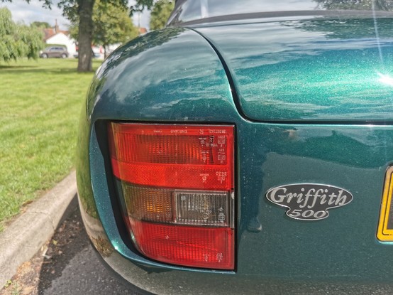 Rear left hand corner of a metallic green TVR Griffith 500. An oval black badge with silver script and edging bearing the model name is visible in the centre of the image.