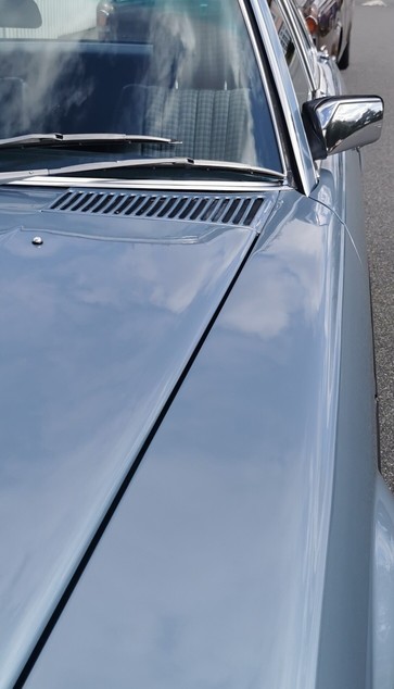 View of the top of the wing and part of the bonnet of a pale metallic blue car.
The windscreen has a chrome surround an the mirror on the passanger door is quite square and chrome backed.
It seems both of the wipers park well over to the passanger side.