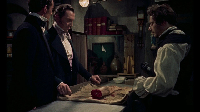 Image du film, trois hommes debout autour d'une table sur laquelle se trouve un bras coupé.