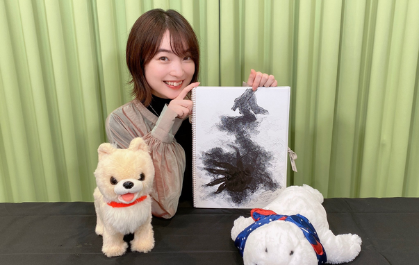 Reina smiles as she shows off a sketch of something with a barking dog and seal plush on the table.