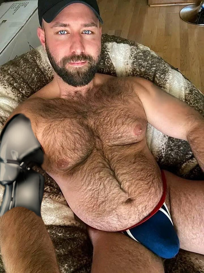 A hairy bear android wearing a black ball cap with a beard and hairy chest has its right forearm torn open revealing white plastic robotics underneath
