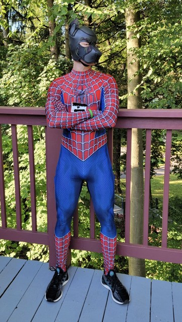 A human dog in cameo hood and Spider-Man costume in front of woods. He crosses his arms and looks into the lens with a smirk