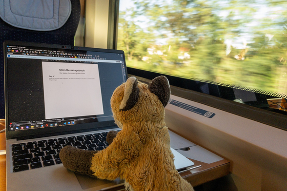 A red fox plushy sitting on a train and writing a travel diary on a laptop