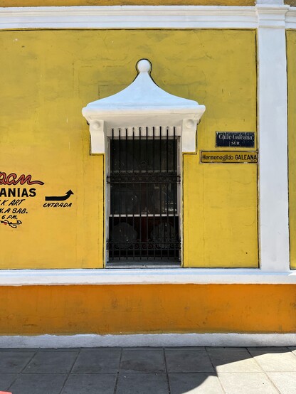 A wall painted yellow and orange is home to a window at the corner of Calle Galeana. The house is an old colonial-style building typical of northern Mexico. Painted on the wall we see part of a sign announcing a fine crafts market with an arrow pointing to the corner to signal the entrance.