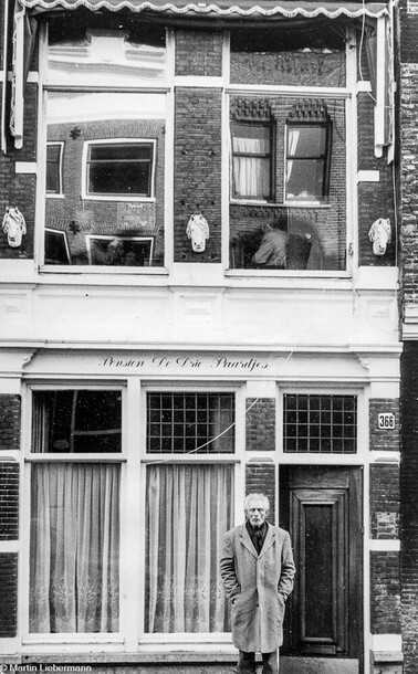 Black and white print in portrait orientation, showing an old pension in Amsterdam. Written on the wall is the name, "Pension De Drie Pardjes". 
The house is very narrow, with two levels. Nearby buildings are reflected in the first floor windows, showing some distortions. A man and a white horses head are dimly visible through one window. Three sculptures of three white horses heads jut from the brick wall between the windows. A thin old man with a white beard is standing before the wooden entrance door. His eyes are closed, his hands shoved into a light grey winter coat. He is standing between the door and two tall windows with long curtains.
