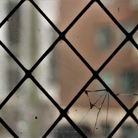 A close up image of a dirty leaded glass window. The bottom right portion has cracks spreading through the section. Despite the dirt and blur, it is possible to make out the outline of a red brick house through the window.
