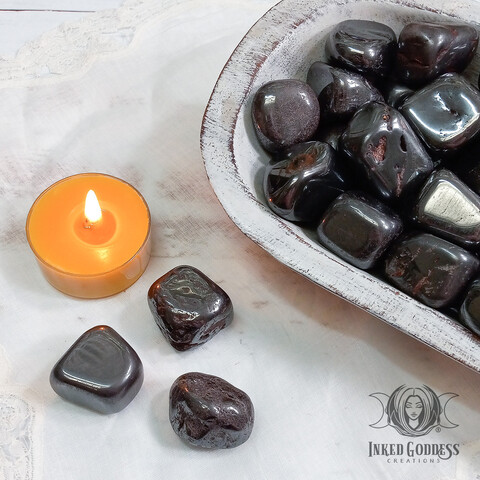 A photo of our "Hematite Tumbled Gemstone" product. It features several specimens in a whitewashed wood bowl with three specimens on a white wood tabletop next to the bowl. An orange tealight candle is shown for scale. Presented by Inked Goddess Creations.