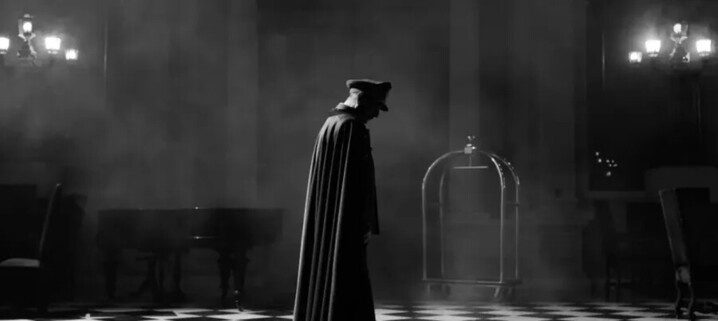 black and white still from the Spanish language film El Conde, depicting the former dictator Augusto Pinochet as a vampire, standing in a gran room, mist or smoke swirling around as he stands in uniform cap and long cape