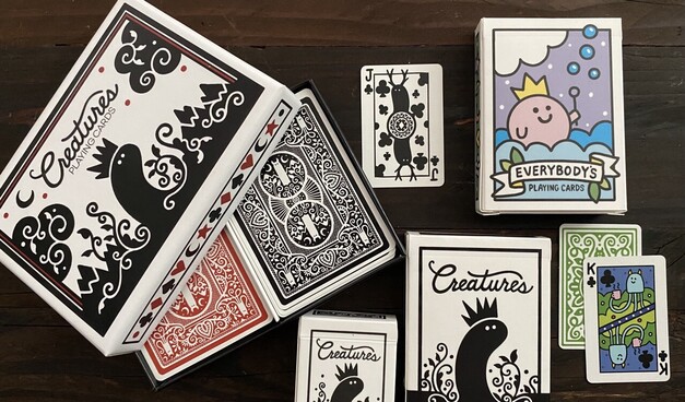 Several decks of playing cards on a table, all in my artwork styles of simple silhouetted creatures. Some are black white and red, others are less traditional and more cartoony.