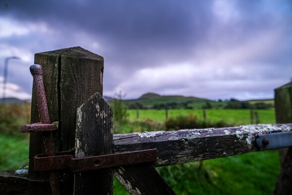 Shot focused on gate post and rusty hitch / catch. Background of farmland with mountains in distance, cloud covered heavy sky above