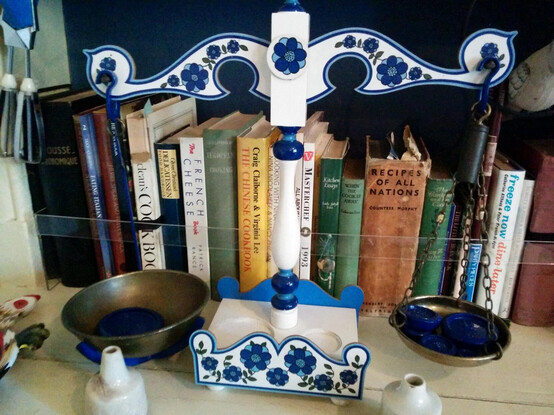 An assortment of hard- and soft-backed cookery books stand in a row filling a shelf. The shelving's white and the background wall is painted deep blue. A clear protective strip runs across the shelf part way up the books. A set of two-pan scales sits in front of the books, it's body looks made of jigsawed wooden pieces painted white with a blue 70s floral design. Two white or cream pie funnels are in front of the scale pans and a blue hand whisk with metal beaters can be seen hanging down from the top left.