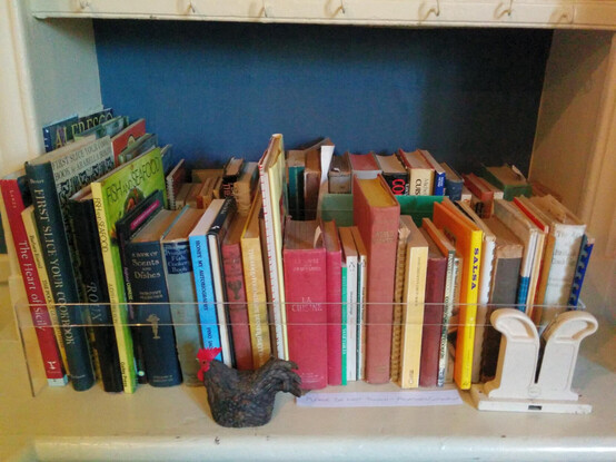 An assortment of hard- and soft-backed cookery books stand in two rows filling a shelf, one row in front of the other. The shelving's white and the background wall is painted deep blue. A clear protective strip runs across the shelf part way up the books and a handwritten sign on white card tells visitors not to touch them. A small black chicken ornament with a red comb and a white object that could be a serviette holder are placed on the shelf on either side of the sign.