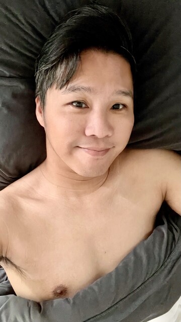 Asian guy laying in bed, selfie with slight smile. His shirtless and half covered under a blanket.