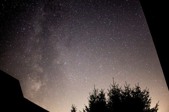 A photo of the Milky Way. The bottom of the photo is glowing slightly orange due to light pollution from the city. Part of a house and part of a tree can be seen as well.