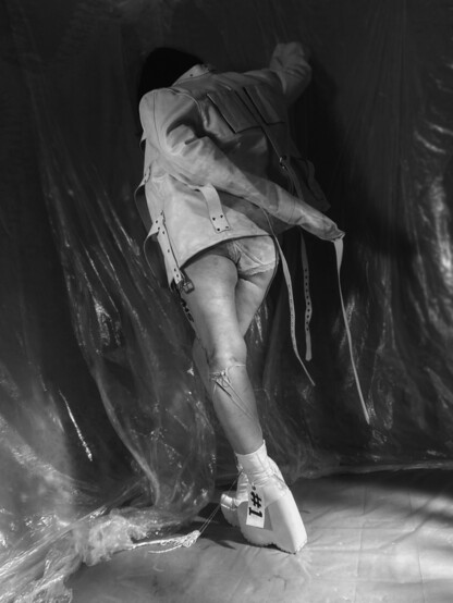 Woman in a room covered in plastic sheets, she's leaning in a corner, wearing white platform boots, white underwear and a white leather straitjacket.