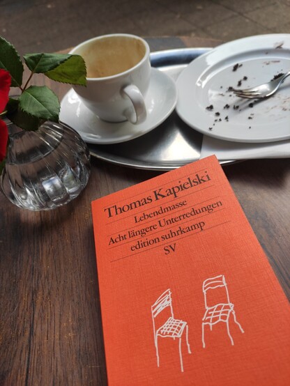 Leere Kaffeetasse, leerer Kuchenteller und ein orngefarbenes Buch, Thomas Kapielski, Lebendmasse.