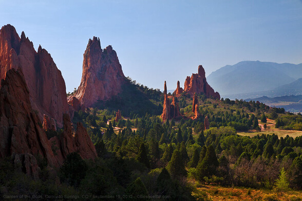 A dramatic landscape of steep, pointed sandstone pinnacles, red in color and half in shade, surrounded by cone shaped evergreens; a tall, round mountain in the distance, back right.
©BosqueBill.com