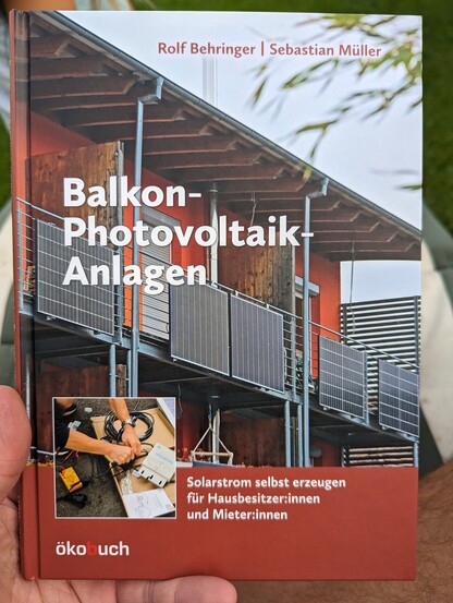 Eine Hand hÃ¤lt ein Buch, auf dem eine Hausfassade mit Solarmodulen sowie ein Werkstatttisch zu sehen sind.
Titel des Buches ist "Balkon-Photovoltaik-Anlagen"