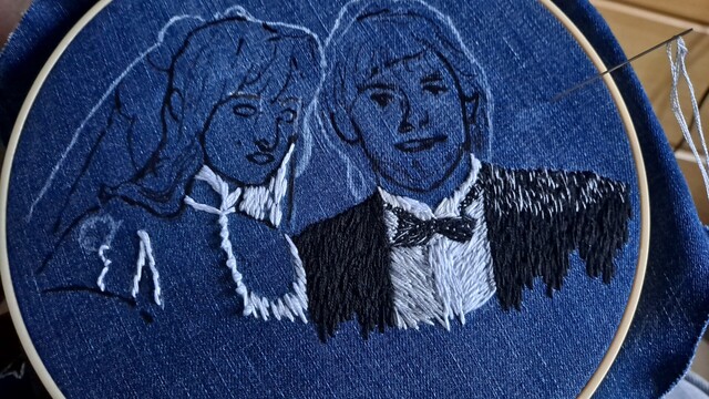 Embroidery in progress on denim in a hoop. Most of it is a sketch depicting a heterosexual couple in wedding garb. The woman's dress has some white outlines stitched in as well as some French knots to show pearl buttons down the front. The man's suit is filled in with shades of black, white, and grey for shadows. His bowtie is a little bit messy, and it's black with white spots. The rest is still just sketches.