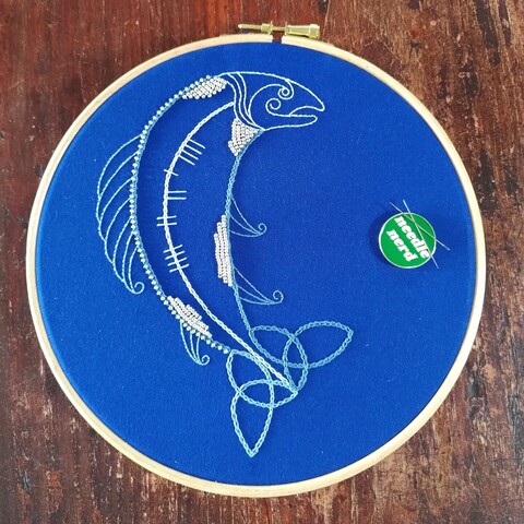 Embroidered salmon in shades of blue thread on blue fabric with silver beads. Ogham writing on the salmon body reads knowledge recalling the Finn McCool legend about the Salmon of Knowledge