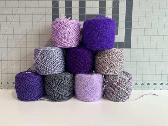 9 wound up yarn cakes stacked in a pyramid shape. The bottom row has a purple, dark grey, a fluffy pink, and speckled light gray and pink yarn. The next row up has a lavendar yarn, a more neon purple yarn, and a rosÃ© colored yarn. The top row has a purple pink yarn, and a neon purple fuzzy yarn