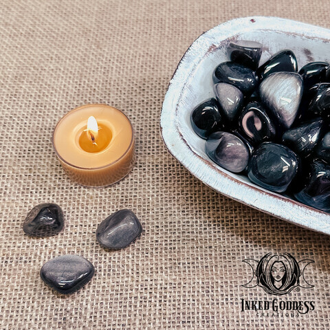 A photo of our "Silver Sheen Obsidian Tumbled Gemstone" product. It features several specimens in a whitewashed wood bowl with three specimens on the burlap-covered surface next to the bowl. A gold tealight candle is shown for scale. Presented by Inked Goddess Creations.