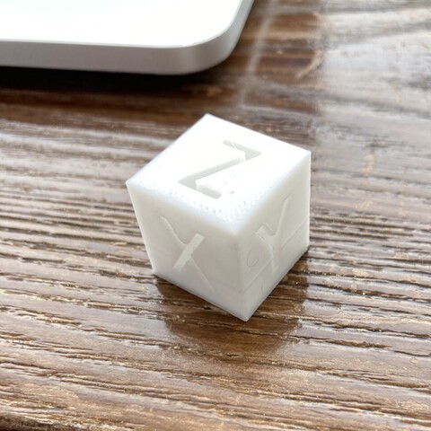 White cube with XYZ on faces. Brown woody grained surface, corner or MacBook in upper left.