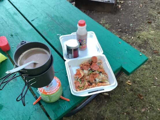 A small backpack cooker with a cup of coffee on the burner is next to a styrofoam food container with pizza on a table top. Moisture from the morning dew is puddled on the top as well