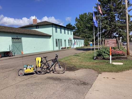 A view of the Great Hinckley Fire Museum in Hinckley MN USA. The museum is a low building with a 2 story central structure, all painted in light treal/green. A bicycle with a trailer is in the foreground perpendicular to the camera view