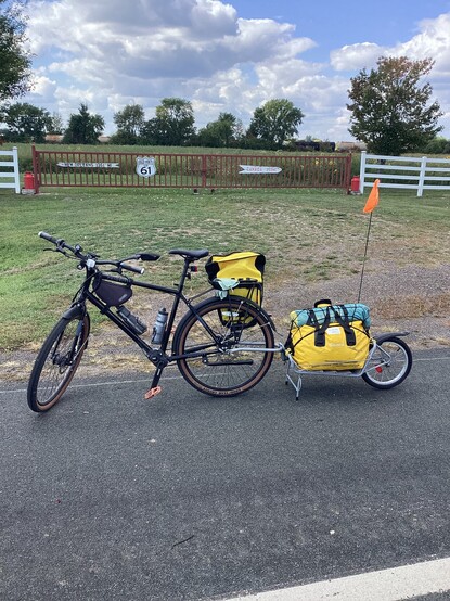 A bicycle with trailer posed in front of a fence where old road signs are hung.  On the left is a sign with a left pointing arrow reading "New Orleans 1361 miles". In the center is a US Highway sign reading "Old Hwy 61". On the right is a right pointing arrow reading "Canada 250 miles".