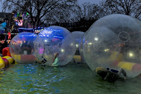 At a fun fair, children roll large plastic bubbles across a body of water. In their midst, a child remains motionless, quietly kneeling at the bottom of his bubble, as if in meditation.