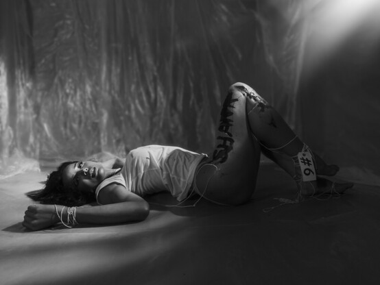 A young woman lies on the floor of a sparse room wearing a white t-shirt, she's loosely tied up with string and has words painted on her legs, hazy light from above shines across her face.