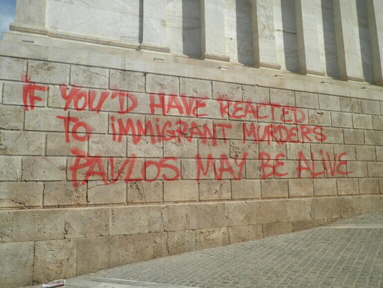 A photograph of a wall with writing in red on it. It reads: IF YOU'D HAVE REACTED TO IMMIGRANT MURDERS PAVLOS MAY BE ALIVE