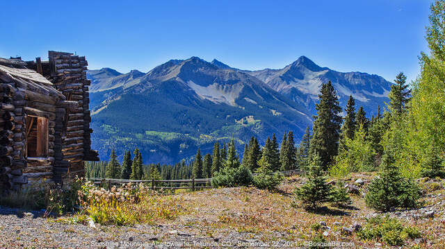 A rugged set of mountains are blue, dark green and green, under a clear blue sky. In the foreground is a sunny clearing bordered by evergreens and, at left, the remains of a wood building.
©BosqueBill.com