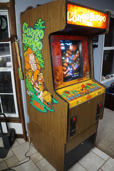 A photo of a Congo Bongo arcade cabinet. It is a standard upright cabinet, with distinct wood panelling. It has "Congo Bongo" illustrations along the sides (showing a giant gorilla and the player character, a man in explorer garb). Its marquee says "Congo Bongo" in big letters, and it has a yellow control area.