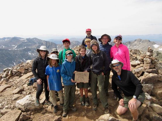 Twelve people huddling together on a high mountain peak holding a sign saying "Quandary Peak 14265"