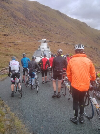 10+ cyclists walking their bikes past a helicopter parked in the middle of the mountain road; behind it is what the english consider to be a mountain