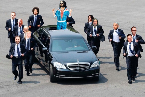Russel Brand stands in an open top limousine, dress in a Jimmy Saville shell suit,  on an airport concreted area, as if just landed receiving his fans, with a security entourage dressed like the men in black, they are all rich, anti woke, rich wing popularists.