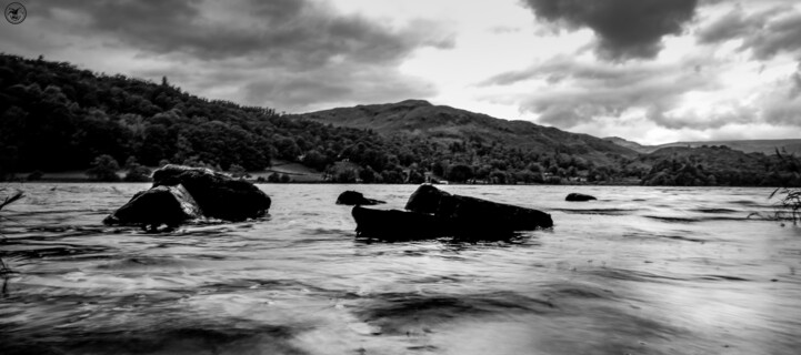 Monochrome shot over lake with underexposed rocks in foreground, mountains and forest on distant shore and cloud covered skies