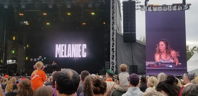 Photograph of a concert stage showing Melanie C on stage in the distance DJing. On one side is a larger projection of the Spice Girl and in the foreground is a little girl wearing an orange shirt with bubbles floating around her head.