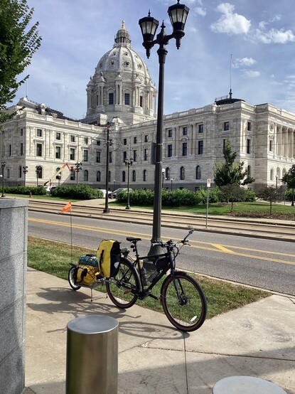 A wide view of the State Capitol of Minnesota USA can be seen across the street behind a bicycle posed in the foreground.  Two wings of the 3 storied while marble can be seen, with the central dome rising above angle where the wings meet.  A blue sky with puffy clouds is above.  All of the doors, windows, and dome have carved figures and scenes decorations