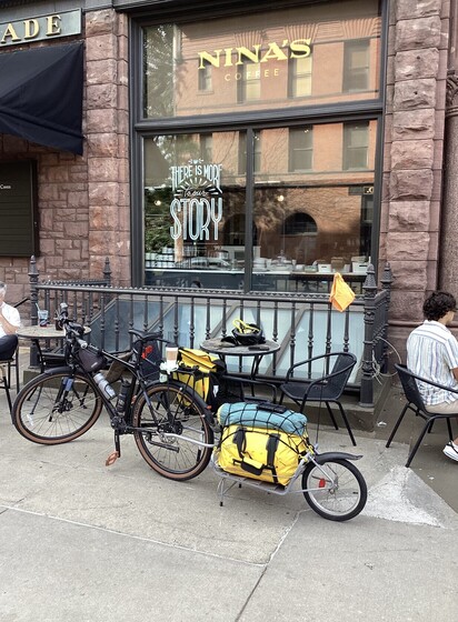 A bicycle with trailer on the sidewalk in front of Nina's Coffee in St Paul, MN, USA with a cup of coffee placed on the seat. A large full story window for the cafe with the name in gold letters is behind.  Small tables and chairs are visible between the bike and building.