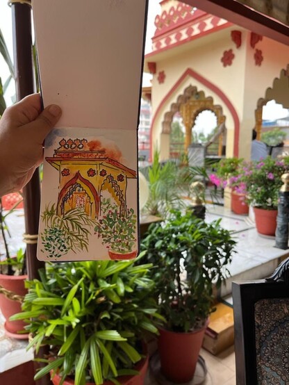 An ink and watercolor sketch of the same Rajasthani canopy in a small cloth bound sketch book. It is being held up by Ruchika next to the subject in the distance.