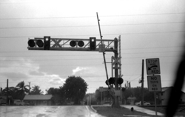 A view through a windshield in heavy rain at a red light. Railroad tracks are ahead with an open gate. There are signs for "railroad crossing - two tracks", "no turn on red", and "stop here at red".