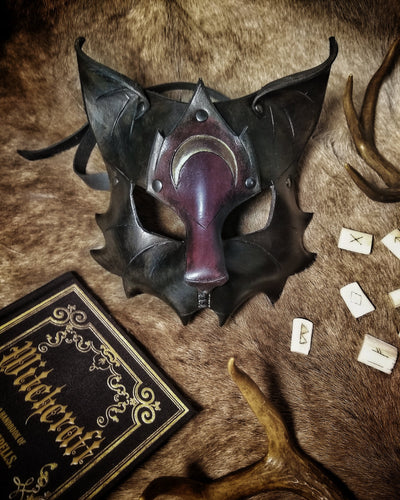 Photo of an intricately detailed leather half-face mask depicting a cat, it has an muzzle extending out from the face, pointed-back ears, a half-moon rune detailed onto the forehead and fur tuft patterning along the lower edges
