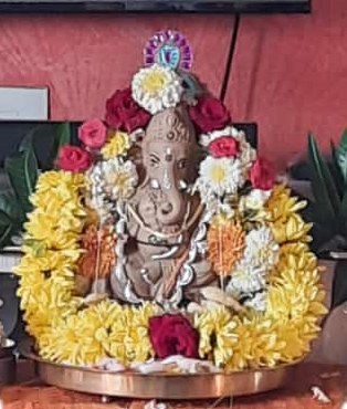 A light brown clay figure of Ganesha decorated with garlands of yellow mums, some white mums around the shoulder, red roses above the white mums, and a red rose at the feet.