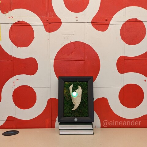 A black frame with a 3D printed section of an animal skull and glowing green eye sits in front of a large red and white logo of Pumping Station: One that is painted on an industrial metal door.