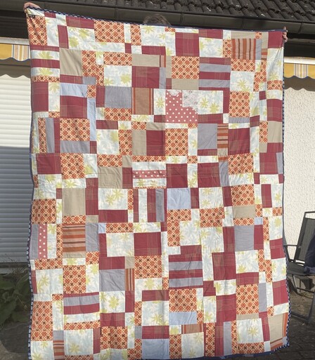 A patchwork quilt made of different prints in orange, dark red and pale blue. It's entirely squares and rectangles, except from one block made in the shape of a cat. The cat is dark pink with white polka dots.